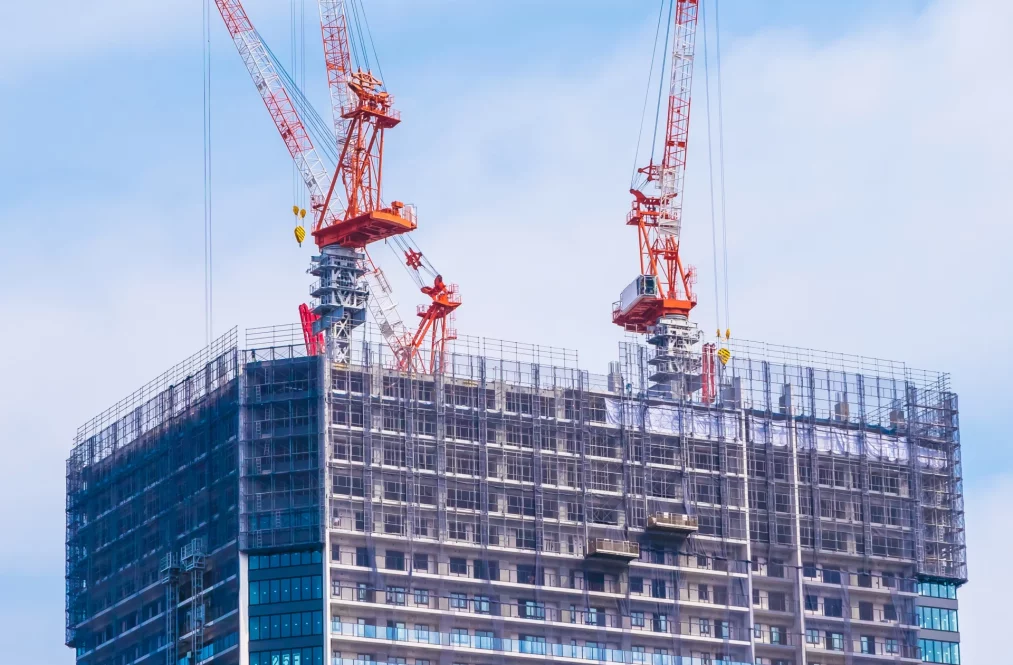 Imagen del proceso de una obra, se pueden ver dos grandes grúas rojas en la cima de un edificio.